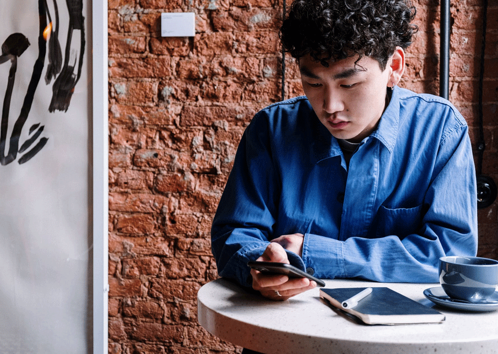 A millennial sits at a round coffee table on his phone at a table with coffee and a notebook. There is a brick background behind him.