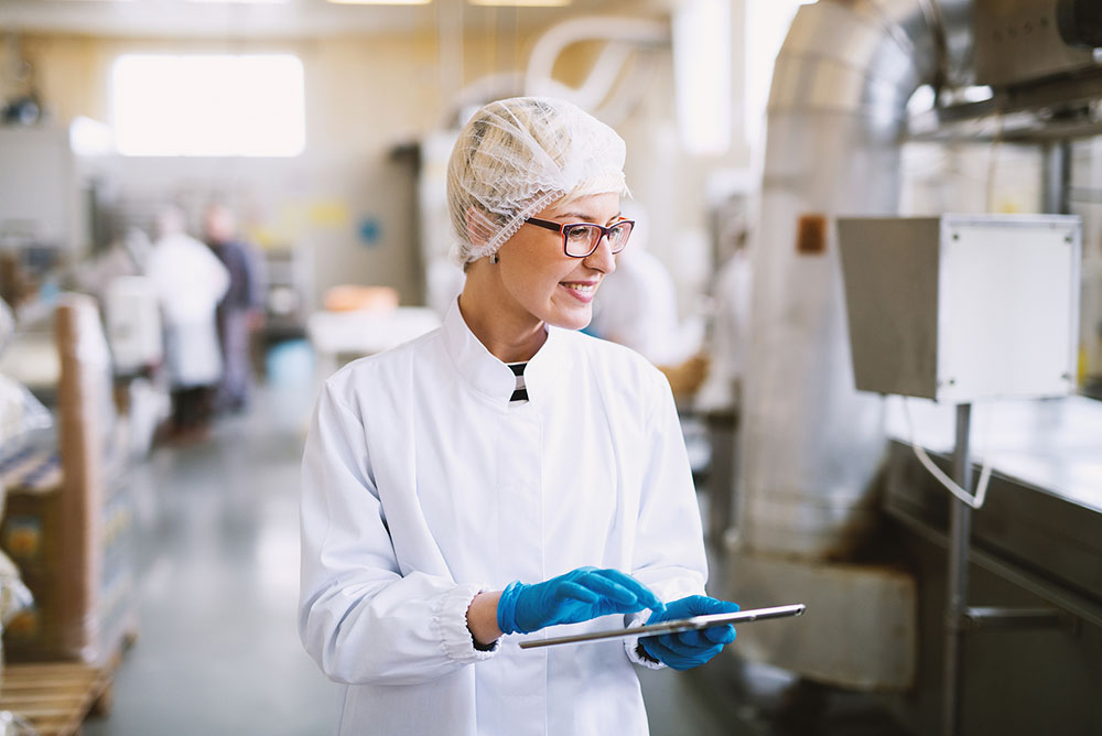 Smiling female worker with glasses in sterile clothes Leveraging MDM for FSMA Compliance as she is holding a tablet in a food factory checking the line.